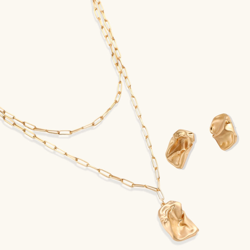 Sculptural gold-toned jewelry set with organic fluid shapes including earrings and layered chain necklace on cream background.
