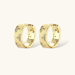 Gold-plated hoop earrings with textured surface and raised floral motifs set with clear zirconia stones on cream background.