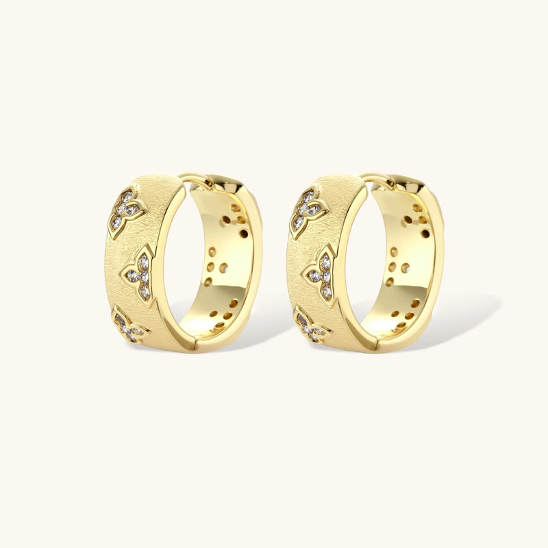 Gold-plated hoop earrings with textured surface and raised floral motifs set with clear zirconia stones on cream background.