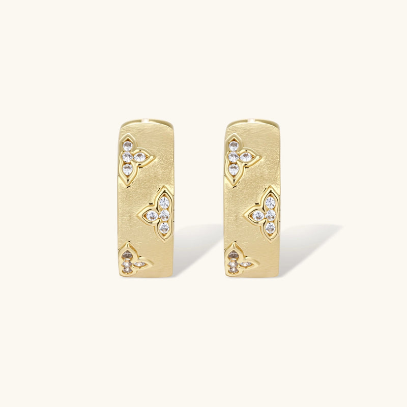 Gold-plated hoop earrings with textured surface and raised floral motifs set with clear zirconia stones on cream background.